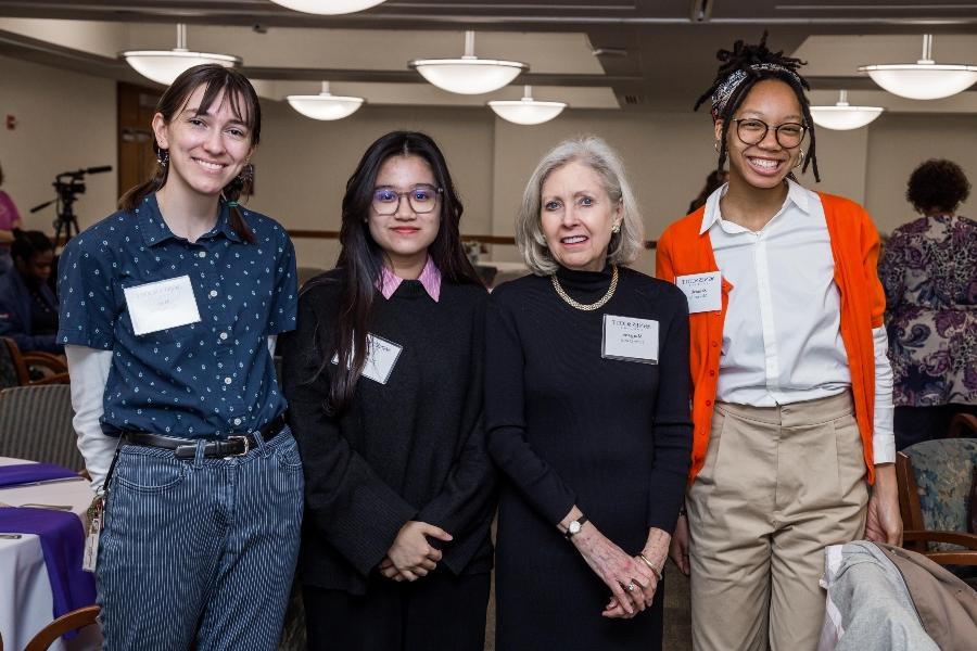 A group photo featuring four women. From left to right, they are standing and smiling, wearing name tags identifying them as participants in the Agnes Scott College event. The woman on the far left is wearing an orange cardigan and glasses, the next woman has short blonde hair and is dressed in black, the third woman is wearing glasses and a black outfit, and the woman on the far right has a blue polka dot shirt and light blue jeans.