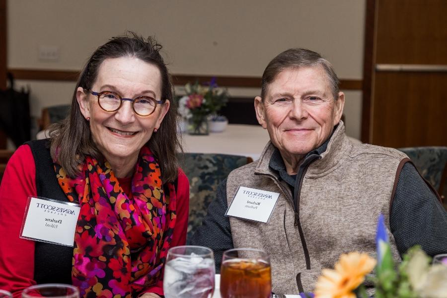 A close-up of two people sitting together at a dining table, both smiling. They are wearing name tags for Agnes Scott College. The man is wearing a beige zip-up vest over a black shirt, and the woman is wearing glasses, a colorful scarf, and a red top.